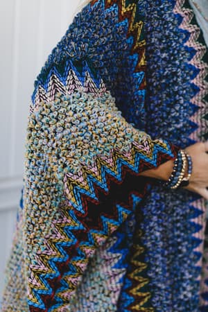 A close-up shows a person's arm in the Desert Dreams Crochet Ruana - Cobalt, with bold zigzag patterns on soft knit fabric. Beaded bracelets add a playful accent, making it an ideal addition to any boho wardrobe.