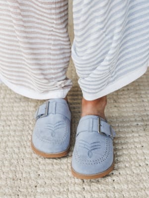 A close-up foot shot of dusty blue suede mules with studded detailing and buckle straps, paired with soft striped wide-leg pants on a chunky woven rug — relaxed and quietly put-together.
