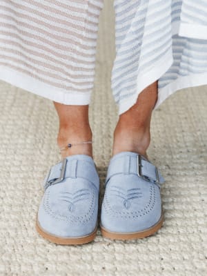 A close-up of periwinkle suede mules with silver buckle hardware and brogue-style stitching detail, styled with a striped knit midi skirt and a delicate evil eye anklet — effortless and a little personal.