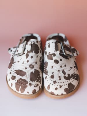 A pair of cow-print mule clogs shot from above against a dusty rose backdrop — cream and brown spots meet silver stud detailing and a buckle strap for a look that's equal parts western and wearable.