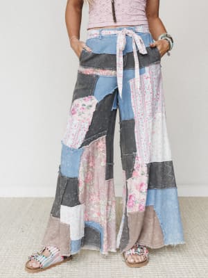 A statement-making pair of ultra-wide-leg patchwork pants stitched together from floral, striped, and washed denim fabrics — worn with a pink crochet crop top and beaded sandals for a free-spirited, artsy-boho vibe.