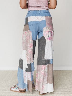 Shot from behind, these wide-leg patchwork pants mix denim, floral prints, and worn fabric panels in dusty pinks and grays — styled with a blush lace top and embellished sandals against a clean studio backdrop.