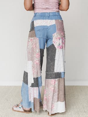 Shot from behind, these wide-leg patchwork pants mix denim, floral prints, and worn fabric panels in dusty pinks and grays — styled with a blush lace top and embellished sandals against a clean studio backdrop.
