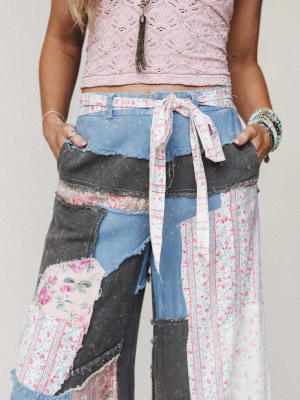 A close-up of patchwork wide-leg denim pants layered with floral and striped fabric panels, styled with a pink crochet crop top and a fabric tie belt — the kind of pants that tell a whole story before you even say a word.