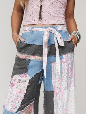 A close-up of patchwork wide-leg denim pants layered with floral and striped fabric panels, styled with a pink crochet crop top and a fabric tie belt — the kind of pants that tell a whole story before you even say a word.