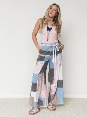 A smiling model in a cropped pink lace tank and show-stopping patchwork wide-leg pants — a mix of denim, floral fabric, and raw-edge seams that feels like your coolest vintage find finally made it into a real pair of pants.