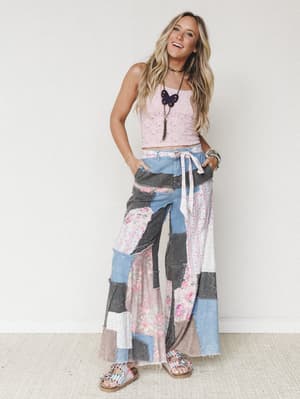 A smiling model in a cropped pink lace tank and show-stopping patchwork wide-leg pants — a mix of denim, floral fabric, and raw-edge seams that feels like your coolest vintage find finally made it into a real pair of pants.