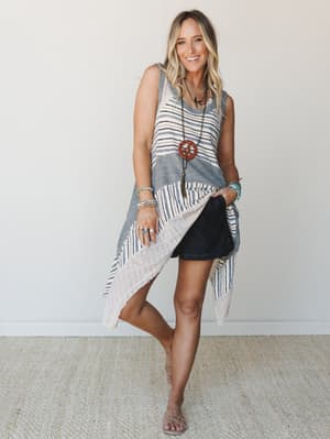A striped knit tunic with a dramatic asymmetric hem is layered over dark denim shorts, styled with layered turquoise jewelry and a peace sign pendant — the kind of effortless warm-weather outfit that looks like you tried just enough.