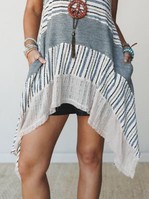 A close-cropped mid-shot showing a patchwork knit tunic with a dramatic asymmetric hem, layered over dark denim shorts — the mixed stripe textures and handkerchief hem give it that effortlessly collected, lived-in boho feel.