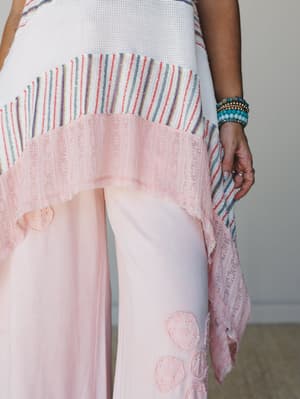 A close-up detail shot of a mixed-media knit tunic layered over wide-leg pink pants, showing a patchwork of waffle knit, multicolor stripe fabric, and a blush lace hem — worn with a stack of turquoise and beaded bracelets that feel like they've been