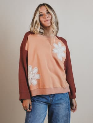 Woman wearing a peach sweater with white floral designs and a brown cardigan against a plain background
