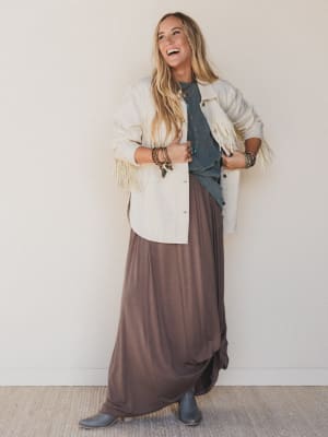 Woman wearing a white cardigan with fringe, brown skirt, and blue top against a white wall.