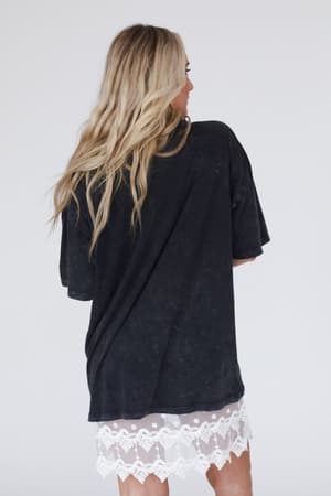 A woman with long, wavy blonde hair stands facing away, wearing the Share The Love Graphic Tee in charcoal over a white lace-trimmed skirt against a plain light background.