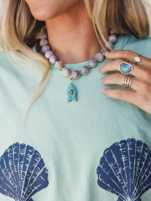 A close-up styled shot of a chunky lavender-grey beaded necklace with a turquoise fish charm, layered over a mint tee printed with navy scallop shells — the kind of effortless coastal styling that looks like it belongs near salt water.