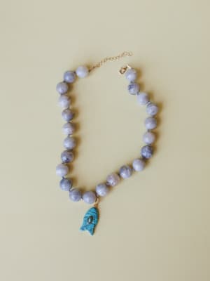 A chunky grey marbled bead necklace laid flat against a warm sage background, anchored by a carved turquoise fish charm — the kind of piece that feels like a souvenir from somewhere you actually want to go back to.