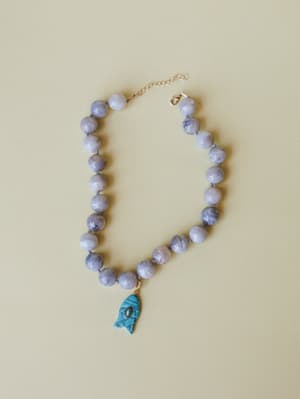 A chunky grey marbled bead necklace laid flat against a warm sage background, anchored by a carved turquoise fish charm — the kind of piece that feels like a souvenir from somewhere you actually want to go back to.