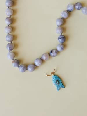 A close-up of a knotted beaded necklace with soft lavender-grey marbled stone beads, finished with a carved turquoise fish charm on a gold hook — the kind of piece that feels like it came home from a coastal antique market.