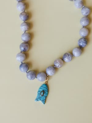 A close-up of a knotted beaded necklace in soft lavender-grey stone beads, centered by a carved turquoise fish charm with gold hardware — the kind of piece that looks like it came home from a coastal market and never left your neck.