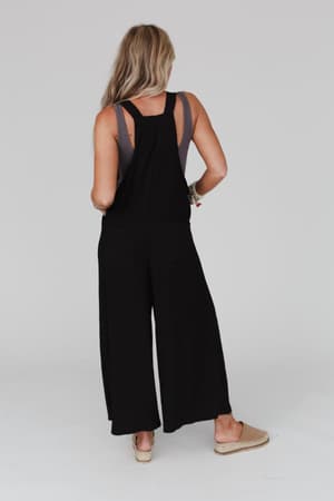 A woman with long blonde hair faces away, wearing The Nest Serenity Wide Leg Jumpsuit - Black over a gray tank top. She has one hand on her hip and beige slip-on shoes. The background is plain and light-colored.