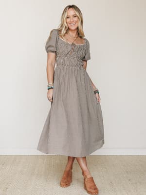 A gingham midi dress with puff sleeves and a gathered skirt styled with tan clogs and stacked turquoise bracelets — the kind of outfit that feels equally at home at a farmers market or a backyard birthday party.