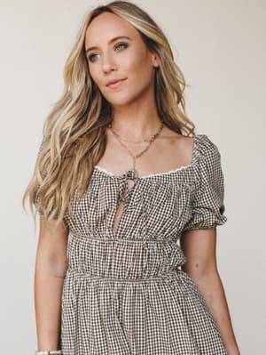 A gingham dress in warm brown and cream does all the talking here — the tie-front neckline and lace trim give it that sweet, lived-in cottagecore feel, styled with layered gold necklaces for just enough polish.