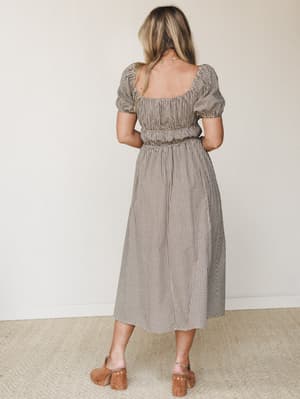 A back view of a brown gingham midi dress with puff sleeves and a cinched elastic waist, styled with tan block-heel mules — the kind of dress that feels equally at home at a farmers market or a summer birthday dinner.