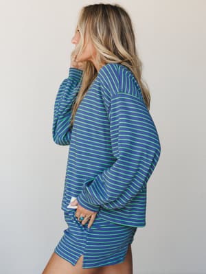A side-profile shot of a model in a matching ribbed stripe set — bold cobalt and neon green horizontal stripes give this relaxed long-sleeve top a retro-sporty energy, styled with a coordinating mini skirt and stacked turquoise rings.