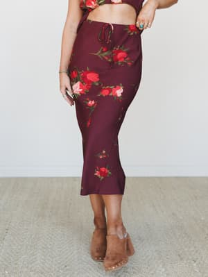 A flowy midi skirt in deep wine with bold red and blush floral print, styled as a matching set with a cropped top and paired with tan studded clogs — romantic and a little vintage in the best way.