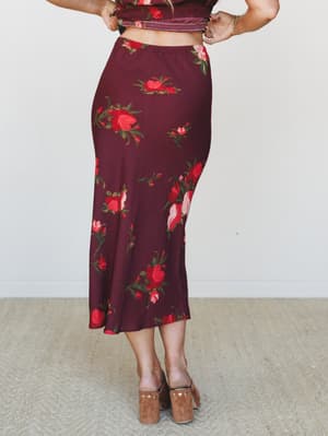 A back-facing model shows off a wine-toned floral midi skirt with a fluid, bias-cut silhouette, styled with a matching smocked crop top and tan suede block-heel mules — warm, romantic, and made for fall.