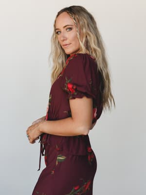 A side-profile shot of a model in a deep wine floral blouse with puff sleeves and a tie waist, paired with a matching floral skirt or pant — romantic and effortless against a soft neutral studio backdrop.