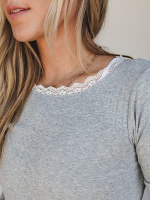 A close-up of a heather grey pointelle knit top with delicate white lace trim at the neckline, layered with fine gold chain necklaces — soft, feminine, and the kind of top you reach for on days you want to feel put-together without trying.