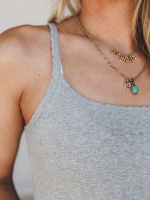A close-up detail shot of a heather grey pointelle cami with delicate lettuce-edge trim, styled with layered gold charm necklaces including a turquoise stone pendant — soft, personal, and quietly pretty.