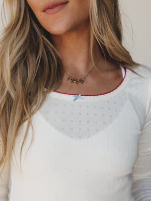 A close-up detail shot of a cream pointelle long-sleeve top with a red contrast trim neckline and a small blue bow accent, styled with a gold 'MAMA' letter necklace — sweet, personal, and quietly nostalgic.
