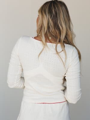 A back-facing shot of a cream pointelle long-sleeve top with delicate red trim detailing at the neckline and hem, styled with a matching cream skirt — soft, understated, and quietly sweet.
