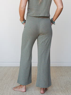 A back-facing model shows off a wide-leg striped pant in warm tan with teal stripe detailing, styled as a matching set and layered with stacked boho bracelets — the kind of outfit you'd wear all day without thinking twice.