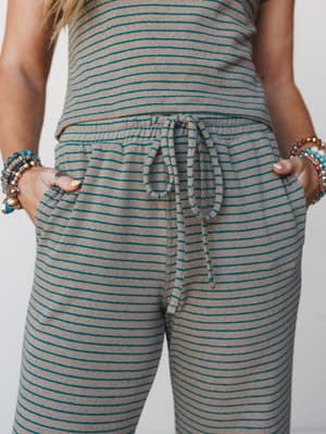 A close-up torso shot of a matching striped set — soft ribbed knit pants with a relaxed drawstring waist, stacked beaded bracelets on both wrists, hands tucked casually in pockets — the whole thing feels like your most comfortable Saturday with a lit