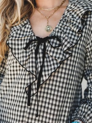 A close-up detail shot of a black and cream gingham top with cascading ruffles and a velvet tie closure, styled with layered gold necklaces and a turquoise bracelet — the kind of top that makes a casual outfit feel like a whole moment.