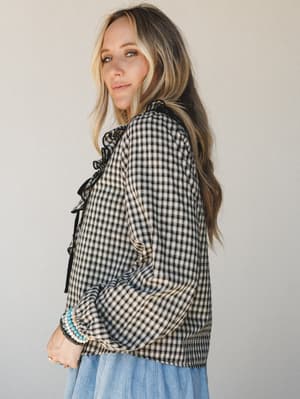 A gingham blouse with ruffle trim and a ribbon tie at the neck, styled over a breezy blue skirt — the kind of top that works just as hard at a farmers market as it does at brunch.