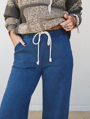 A close mid-shot of wide-leg knit pants in a deep indigo wash, styled with a cropped paisley hoodie and layered boho jewelry — the kind of outfit that blurs the line between staying in and going out.