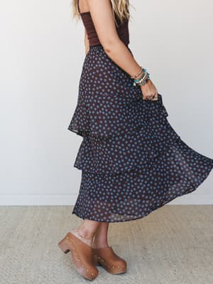 A tiered polka dot midi skirt in deep brown and dusty teal is caught mid-twirl, styled with a chocolate camisole, wooden clogs, and a stacked bracelet arm — effortlessly boho and a little vintage-sweet.