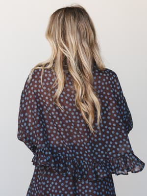 A back-view shot of a flowy polka dot blouse in deep brown and dusty blue, with ruffle hem and cuff details that give it a soft, vintage-feminine feel.