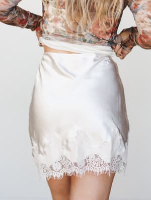 Shot from behind, a satin mini skirt in pearl white with a delicate lace hem pairs with a sheer floral long-sleeve top — the kind of outfit that lives somewhere between a bridal shower and a really good Saturday.