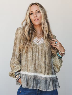 A flowy boho blouse in muted mustard olive with an all-over floral print, lace neckline detail, and a contrast ruffle hem — styled with stacked beaded bracelets and layered gold necklaces for that effortlessly collected, lived-in feel.