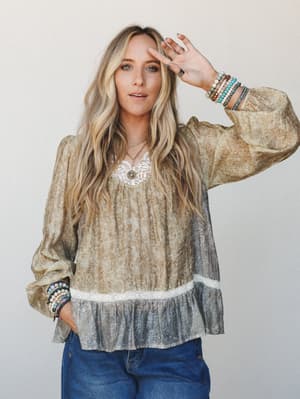 A relaxed boho blouse in mustard olive with a subtle floral print, lace trim neckline and hem band, and a contrast dusty grey ruffle at the bottom — styled with stacked beaded bracelets and a delicate layered necklace for that effortlessly collected