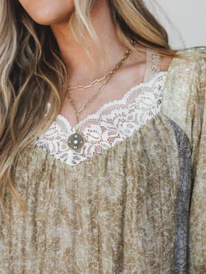 A close-up of a flowy floral blouse in muted olive with delicate white lace trim at the neckline, layered gold chain necklaces adding just the right amount of warmth — the kind of top that looks like it belongs on a long weekend somewhere pretty.