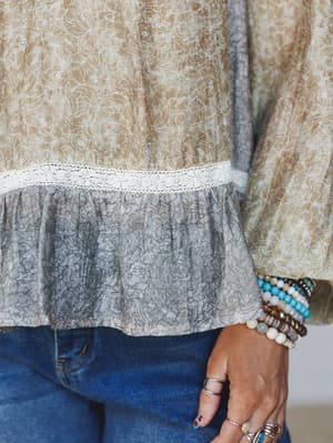A close-up detail shot of a flowy mixed-print blouse with a lace trim waist seam and ruffle hem, styled with stacked beaded bracelets, silver rings, and dark denim — the kind of effortless layering that feels intentional without trying too hard.