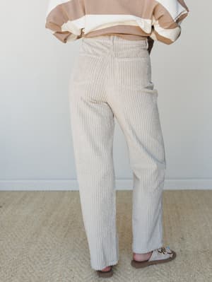 A back-view shot of straight-leg corduroy pants in a soft sandstone tone, styled with a color-block sweatshirt and sandals — the kind of easy, put-together outfit that works from a coffee run to a casual Friday.