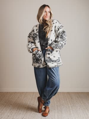 Woman wearing a patterned coat, dark shirt, jeans, and brown shoes against a plain background