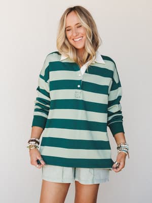 A smiling model wears an oversized rugby-stripe polo top in hunter green and mint paired with light wash shorts, layered bracelets stacked on both wrists — the kind of effortless weekend look that feels like it picked itself.