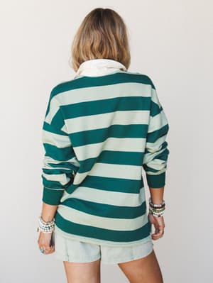 A relaxed back-facing shot of a bold hunter green and sage stripe rugby-style top layered over a cream collared shirt, paired with light denim shorts and stacked beaded bracelets — effortlessly casual with a vintage sports-inspired feel.
